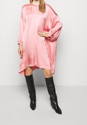 Femme portant une robe ample en satin rose brillant à manches longues et de hautes bottes en cuir noir, debout devant un fond uni.