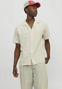 Camisa de manga corta, con botones, en beige claro con un patrón de cuadrícula. Combinada con pantalones de mezclilla desgastados en azul claro. Gorro rosa llevado al revés.
