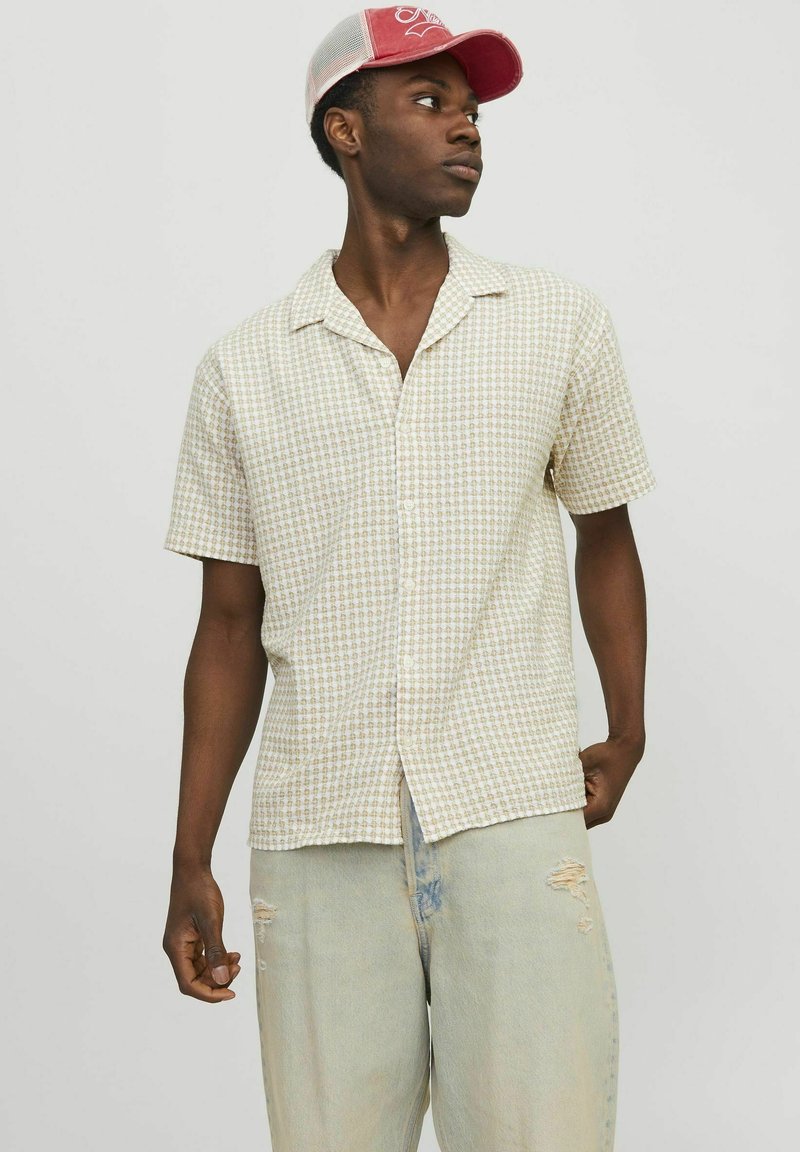 Camisa de manga corta, con botones, en beige claro con un patrón de cuadrícula. Combinada con pantalones de mezclilla desgastados en azul claro. Gorro rosa llevado al revés.