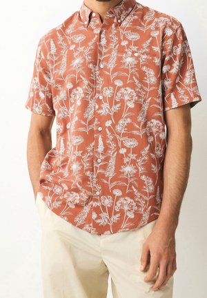 Man wearing rust-orange short-sleeve button-up shirt with white floral pattern and beige pants, hands relaxed by sides.