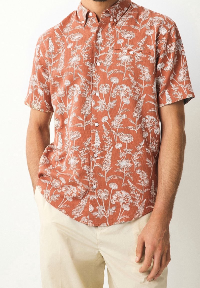 Homme portant une chemise à manches courtes orange rouille avec motif floral blanc et pantalon beige, mains détendues le long du corps.
