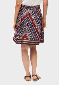 Femme portant une jupe jusqu'aux genoux avec des motifs géométriques colorés, un haut blanc et des sandales plates noires, debout avec le dos tourné vers l'avant.