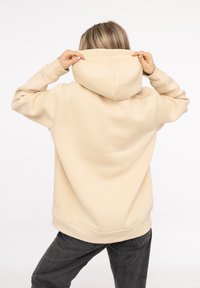 Sudadera con capucha de color beige claro, de corte relajado, textura suave y puños y dobladillo acanalados. La vista trasera muestra la capucha y las costuras.