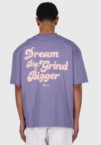Lilla bomulds t-shirt med stor pink tekst på ryggen: "Dream Big, Grind Bigger" med brandnavnet "Dropsize" nedenunder.