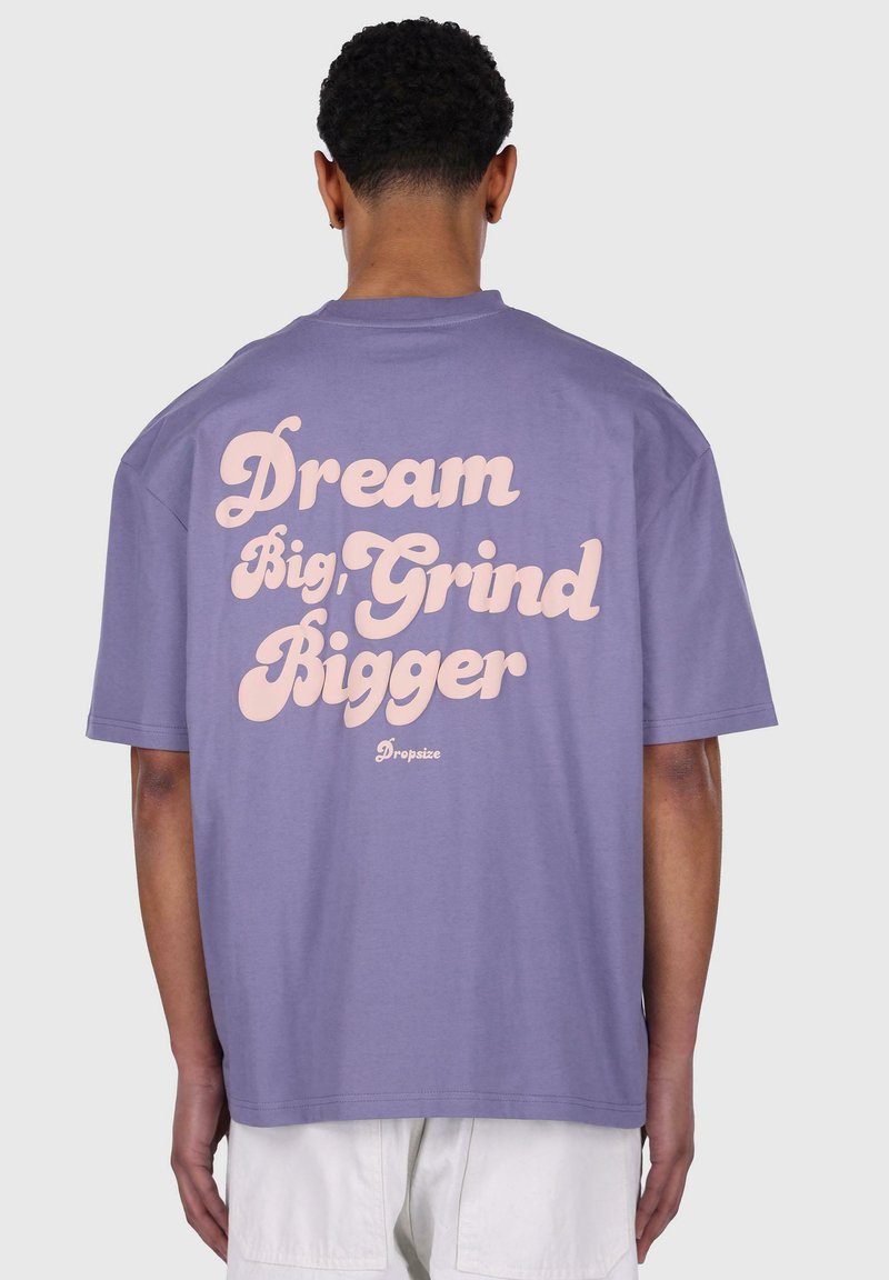 Lilla bomulds t-shirt med stor pink tekst på ryggen: "Dream Big, Grind Bigger" med brandnavnet "Dropsize" nedenunder.