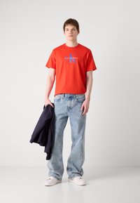 Červené bavlnené tričko s modrým logom "Calvin Klein", v kombinácii s voľnými svetlomodrými džínsy a bielymi teniskami. Tmavý kabát prehodený cez ruku.
