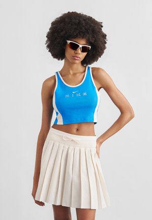 Femme aux cheveux afro portant des lunettes de sport blanches, un crop top bleu Nike et une jupe plissée blanche, posant avec une main sur la hanche.