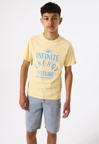 T-shirt en coton jaune avec le texte bleu "ÉNERGIE INFINIE AVENIR" et un graphique de soleil, assortie à un short en denim bleu délavé.