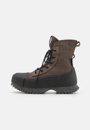 Bota robusta marrón y negra de caña alta con cordones negros, puntera reforzada, suela gruesa texturizada y tiradores de tela en el talón y la lengüeta.