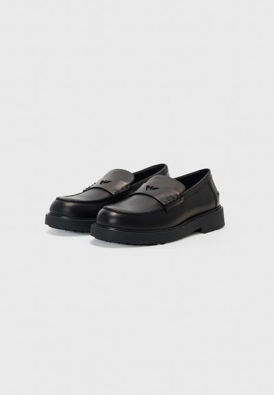 Schwarze Lederloafer mit glatter Oberfläche, leicht erhöhter Gummisohle und genähten Details am Schaft. Subtiles Logo auf dem Riemen.
