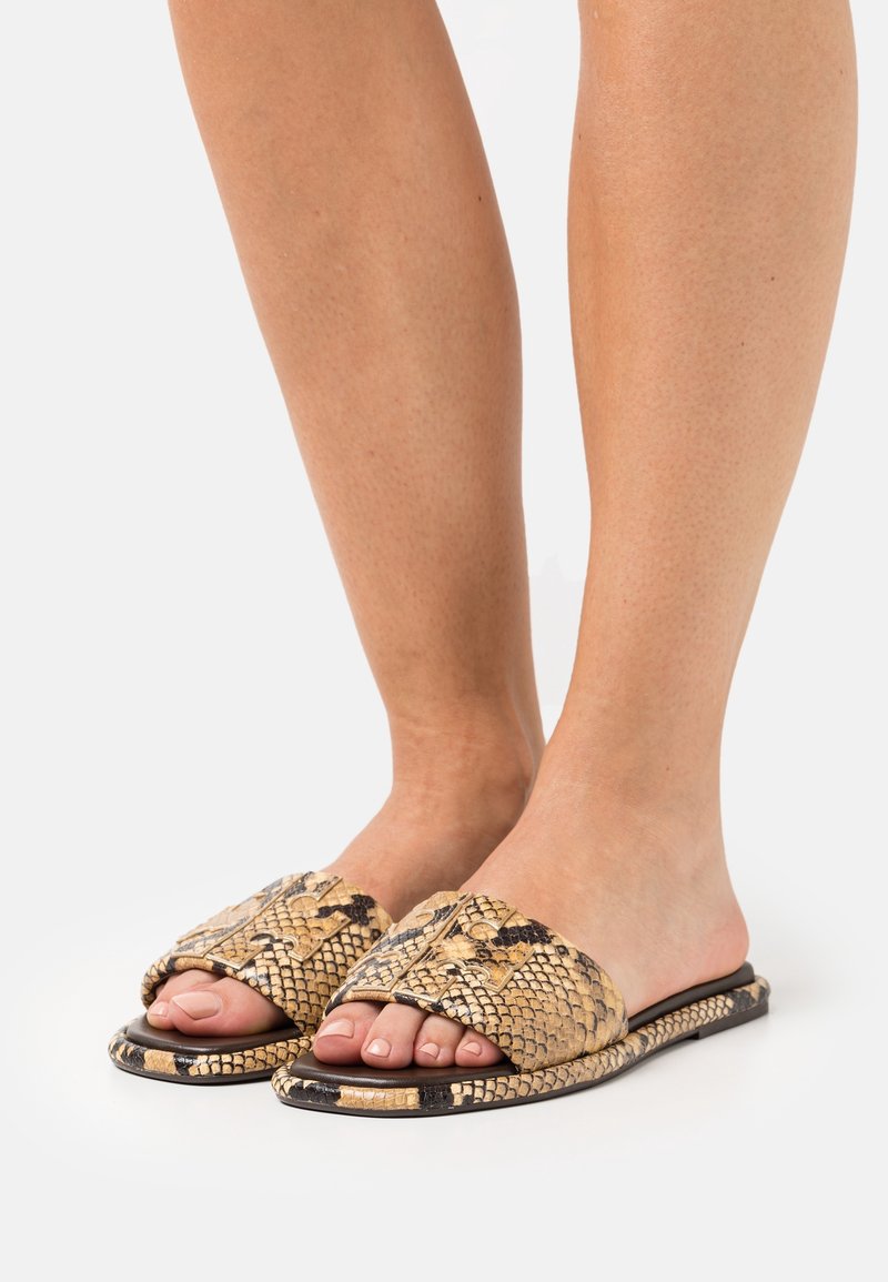 Pieds portant des sandales plates ouvertes à bouts ouverts avec un motif peau de serpent beige et noir sur les brides et les semelles, sur un fond uni.