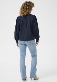 Navyblauwe blouse met lange mouwen en pofschouders, met een plooidetail aan de achterkant, gecombineerd met lichtblauwe flared jeans en beige hakken.