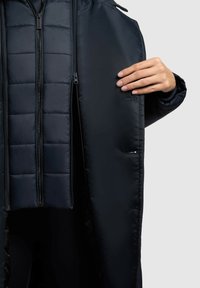 Daunenjacke, schwarz, mit einem gesteppten Innenfutter, mehreren Reißverschlüssen und einer glatten Außenseite. Mit seitlichen Taschen und einer Kapuze.