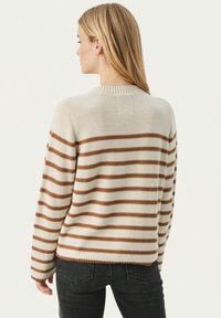 Frau mit langen blonden Haaren, die einen cremefarbenen Pullover mit braunen horizontalen Streifen und dunklen Jeans trägt, steht mit dem Rücken zu einem weißen Hintergrund.