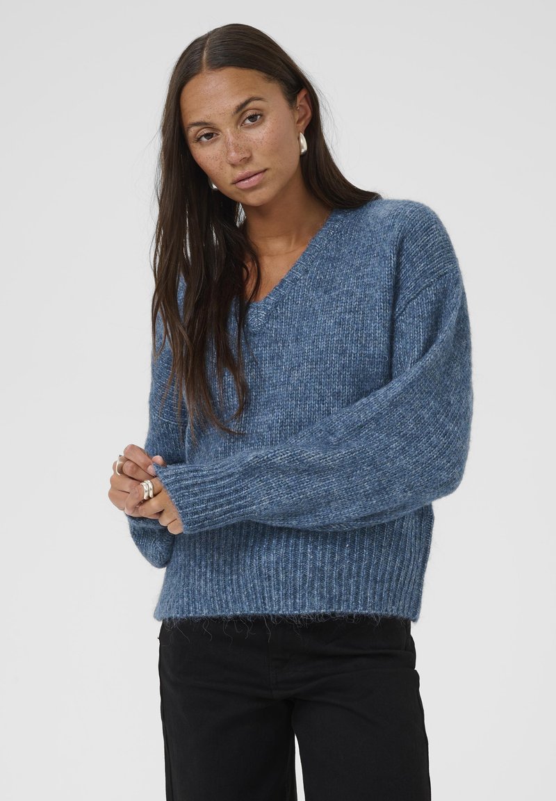 Maglione blu lavorato a maglia con scollatura a V, polsini e orlo a coste. Tessuto morbido con una vestibilità leggermente ampia. Indossato sopra pantaloni neri.