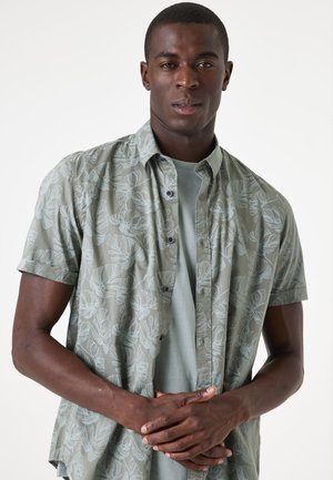 Camisa de botones de manga corta, color verde claro, con un patrón de hojas, superpuesta a una camiseta verde pálido. Presenta botones oscuros y un ajuste relajado.