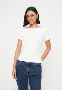 Λευκό πουκάμισο με κοντά μανίκια και ελαφρύ λαιμόκοψη σχήματος βάρκας, με χρυσές λεπτομέρειες στους ώμους και εφαρμοστή γραμμή, συνδυασμένο με μπλε τζιν.