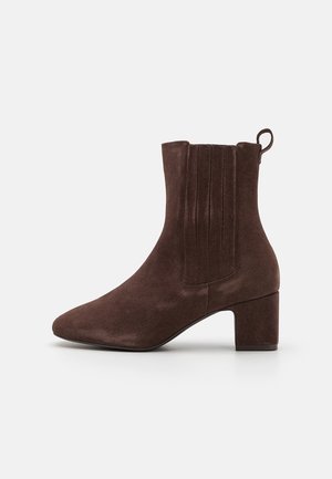 Bottine en daim marron avec un bout pointu, un talon block de hauteur moyenne et des plis verticaux sur le côté extérieur. Dispose d'une languette à l'arrière.