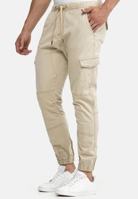 Beige Cargohose mit elastischem Bund und Kordelzug, ausgestattet mit Seitentaschen und elastischen Bündchen, hergestellt aus weichem Baumwollmaterial.