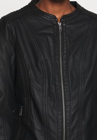 Veste en cuir noir avec un col rond, fermeture éclair, détails matelassés aux épaules et poches latérales. Texture lisse et design ajusté.