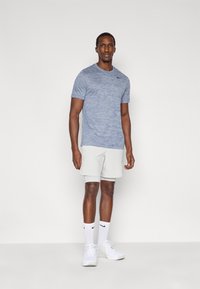 Ανδρικό αθλητικό σύνολο που περιλαμβάνει ένα ελαφρώς σκούρο γαλάζιο κοντομάνικο πουκάμισο και ανοιχτογκρι σορτς, συνδυασμένο με λευκά αθλητικά παπούτσια και κάλτσες αστραγάλου.