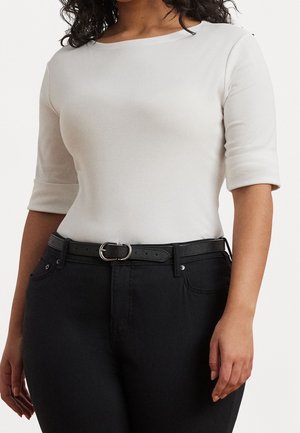 Femme portant un haut blanc ajusté à manches trois-quarts, rentré dans un pantalon noir taille haute avec une ceinture noire.