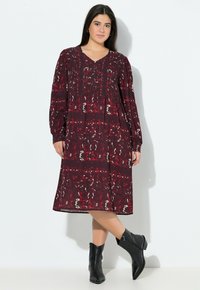 Bordeaux gemustertes Kleid mit langen Ärmeln, einem V-Ausschnitt und fließender Silhouette. Kombiniert mit schwarzen Knöchelstiefeln. Glatte Textur.