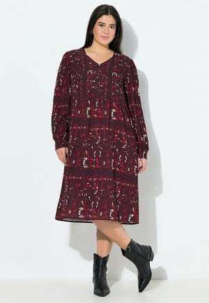 Bordeaux gemustertes Kleid mit langen Ärmeln, einem V-Ausschnitt und fließender Silhouette. Kombiniert mit schwarzen Knöchelstiefeln. Glatte Textur.