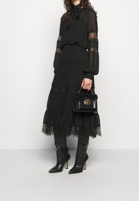 Schwarzes Kleid mit transparenten langen Ärmeln, Spitzenakzenten und gestuftem Rock. Kombiniert mit kniehohen Lederstiefeln und einer kleinen schwarzen Handtasche mit Krokoprägung.