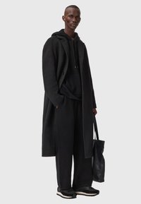 Langer schwarzer Mantel über einem schwarzen Kapuzenpullover und weiten Hosen. Schwarze Schuhe mit weißer Sohle, hält eine strukturierte schwarze Tragetasche.