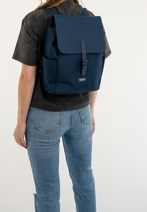 Donkerblauwe rugzak met een bovenklep en gesp sluiting, met een gladde textuur en verstelbare riemen. Minimale merkopdruk aan de voorkant.