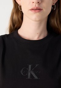 Fekete pamut póló kerek nyakkivágással, amelyen egy diszkrét szürke Calvin Klein logó található a mellkason. Sima textúrájú és testhez simuló dizájn.