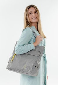 Frau in einem hellblauen Kleid trägt eine große graue, strukturiert Tasche mit gelben Reißverschlussanhängern über der Schulter, lächelt und blickt seitlich.