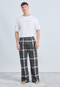 Fehér pamut póló "TOMMY HILFIGER" logóval. Fekete és szürke kockás flanel pizsamanadrág rugalmas derékkal. Modell mezítláb áll.