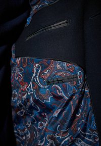 Chaqueta negra con un forro interior de estampado paisley en azul, rojo y blanco, con una textura de tela elegante y dos bolsillos visibles.