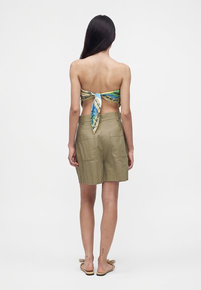 Femme aux longs cheveux foncés portant un top foulard multicolore noué, un short vert olive avec des poches et des sandales plates, debout, vue de dos.