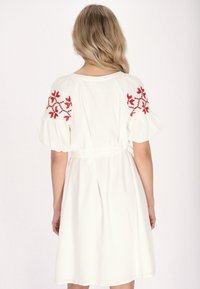 Witte jurk met korte, bolle mouwen en rode bloemenborduring. Het ontwerp heeft een strik in de taille en een vloeiende rok tot de knie.