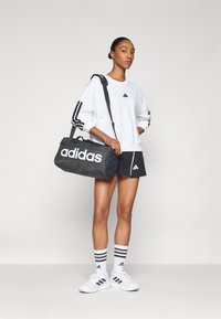 Juodas sportinis krepšys su baltu "adidas" logotipu. Modelis dėvi baltą džemperį su juodomis juostomis, šortus ir baltus sportbačius su juodais akcentais.