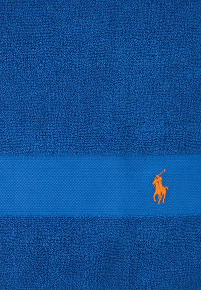 Μπλε πετσέτα με υφή και μια μονή λωρίδα που φέρει ένα μικρό πορτοκαλί λογότυπο με παίκτη πόλο κεντημένο στο κάτω δεξί μέρος.