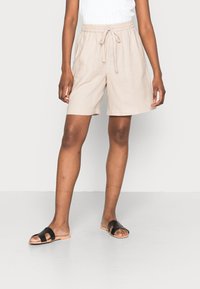 Shorts en lin beige avec une taille élastique et un cordon de serrage. Présentent une coupe ample et une longueur mi-cuisse. Associés à des sandales à glissière noires.