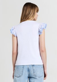 Donna che indossa una maglietta bianca con maniche ruffle ricamate in blu e jeans azzurri, mostrata di spalle su uno sfondo neutro.