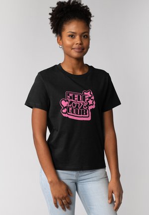 Jonge vrouw met krullend haar draagt een zwart t-shirt met roze "Self Love Club"-opdruk en lichtblauwe spijkerbroek, staand tegen een grijze achtergrond.