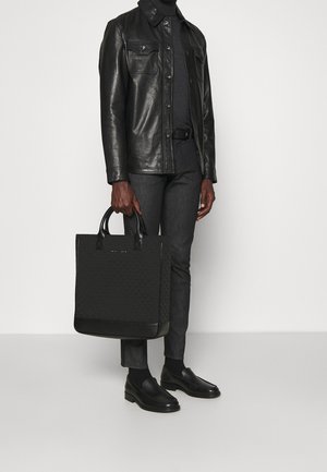 Persoon draagt een zwarte leren jas, zwarte coltrui, slanke zwarte broek, zwarte loafers en houdt een grote zwarte totebag vast.