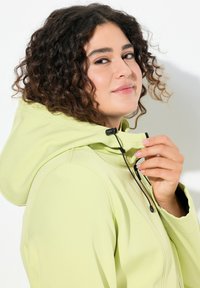 Giacca verde chiaro con cappuccio e texture liscia. Presenta una chiusura con coulisse e zip. Capelli ricci e tonalità di pelle naturali visibili.