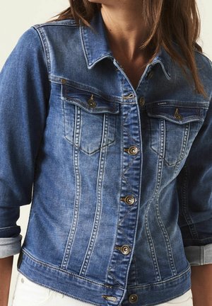 Kobieta w dopasowanej niebieskiej kurtce dżinsowej z podwiniętymi rękawami, zapinanej na guziki z przodu i z dwoma klapkowymi kieszeniami na piersiach.