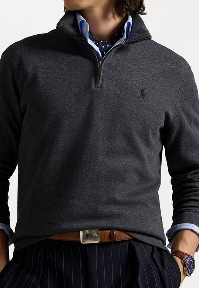Tumši pelēks zip-up pulovers ar teksturētu audumu, ar brūnu rāvējslēdzēju, kārtots virs gaiši zilas krekliņas ar podziņām.
