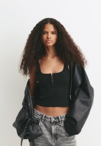 Jonge vrouw met lang krullend haar, gekleed in een zwarte crop top en een oversized leren jas die van haar schouders glijdt, lichte spijkerbroek, neutrale achtergrond.