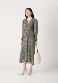 Robe florale à manches longues avec décolleté en V, taille cintrée et jupe fluide dans des tons sombres. Le modèle tient un sac à main crème et porte des talons beiges.