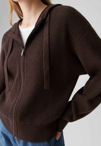 Hoodie marron côtelé avec fermeture éclair, cordons et tissu texturé. Il présente une coupe décontractée et des manches légèrement surdimensionnées.