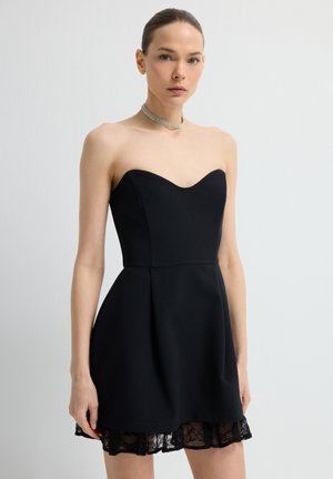 Kvinde iført en sort, stroppløs minikjole med hjerteudskæring og blonder, kombineret med en funklende choker-halskæde.